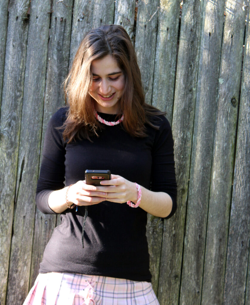 A young woman looking down at her phone. She is wearing a black shirt,and a pink skirt. She has on a spiral superduo necklace in shades of pink and a matching bracelet on her left arm. She is wearing a copper bracelet on her right wrist.