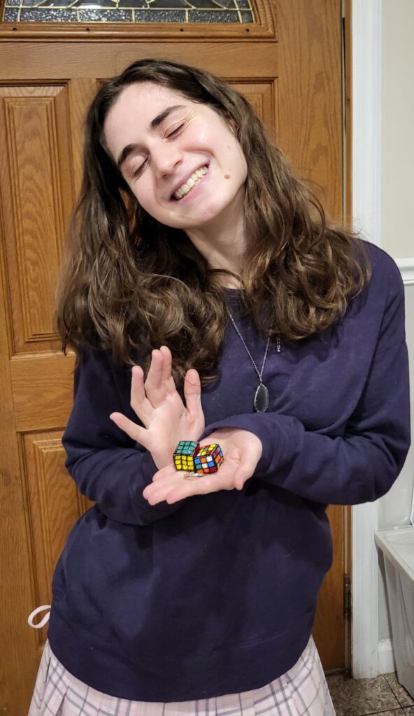 A smiling woman holding two beaded Rubik's Cubes keychains in the palm of her hand. One is "solved" and one isn't.