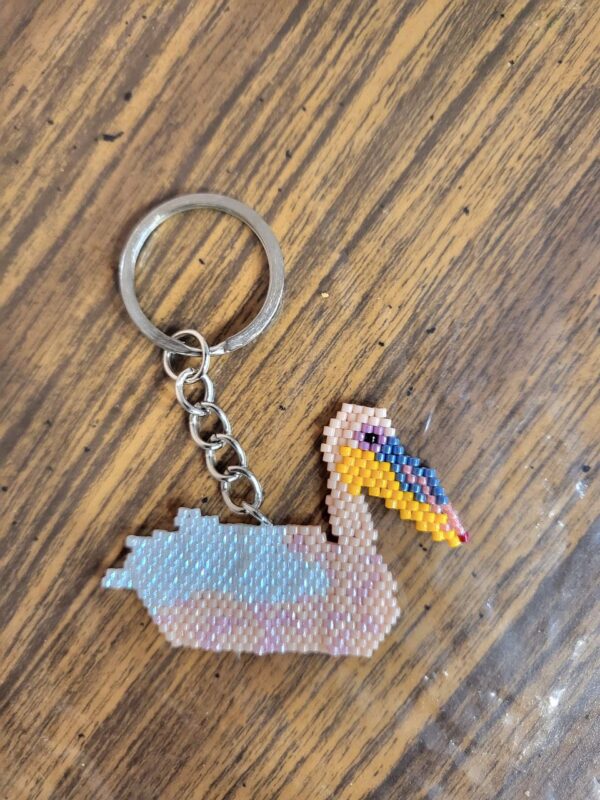 A beaded pelican keychain. The pelican is white and pink and it's on a wooden background.