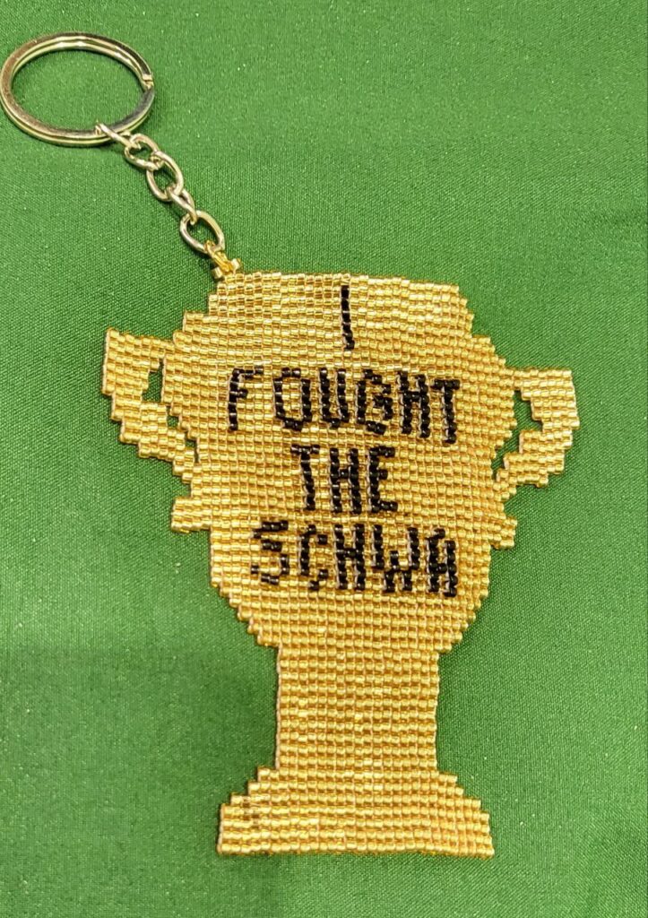 Keychain of a beaded golden trophy with the words "I FOUGHT THE SCHWA" on it in black beads.